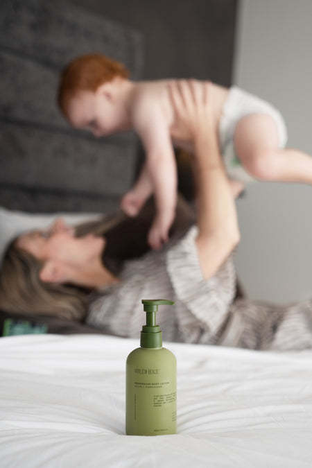 Green bottle Wildheke Body Lotion  on a bed with a blurred background of a person holding a child