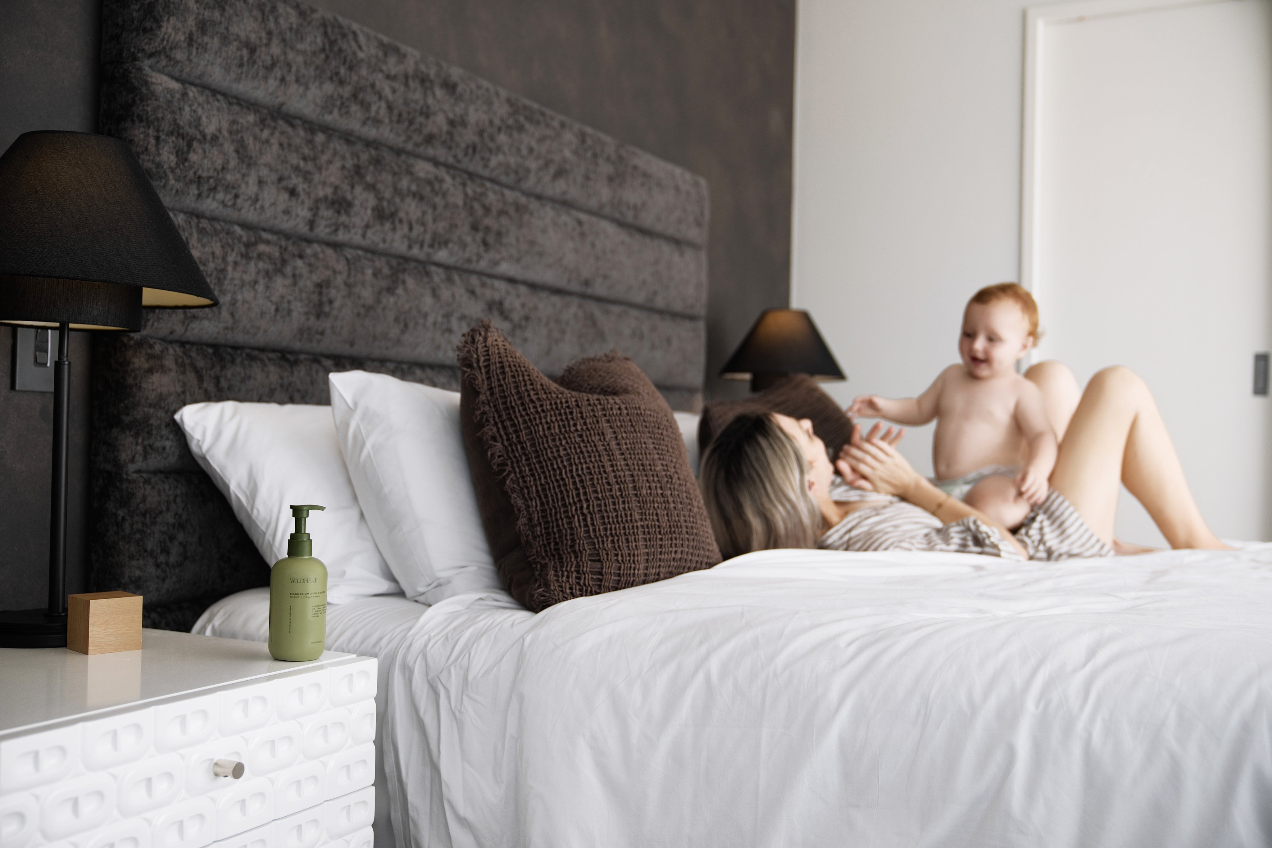 Wildheke Body Lotion - Woman and child on a bed in a modern bedroom with a gray headboard.